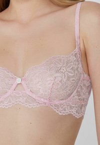 Soutien-gorge en dentelle rose avec des motifs floraux, un détail de nœud en satin au centre, des bretelles réglables et des bords festonnés le long de la bande.