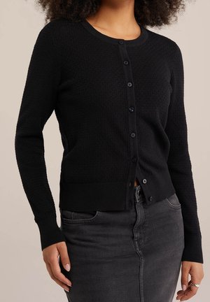 Vrouw die een zwarte textuurvolle, knoopsluitende cardigan met lange mouwen en een donkergrijze spijkerrok draagt, tegen een effen achtergrond.