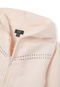 Felpa rosa chiaro con zip, realizzata in tessuto morbido, con borchie decorative argento lungo il petto. Include un cappuccio foderato e un'etichetta interna.