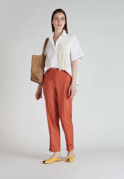 Camicetta bianca a maniche corte con taschino a frange testurizzate, pantaloni su misura color ruggine e mule gialle con punta aperta. Borsa a mano intrecciata marrone.