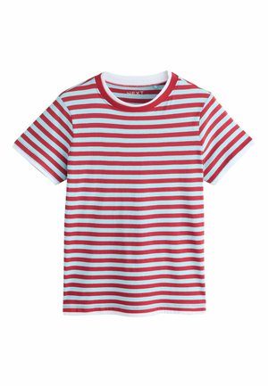 Blonde Frau lehnt an der Wand, trägt ein rot-weiß gestreiftes T-Shirt und einen hellblauen Jeans-Minirock, blickt mit neutralem Gesichtsausdruck nach vorne.