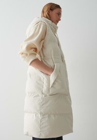 Beige, gepolsterte ärmellose Weste mit Kapuze, seitlichen Taschen und einer glatten, glänzenden Oberfläche, die einen lässigen Schnitt und vertikale Nähte aufweist.