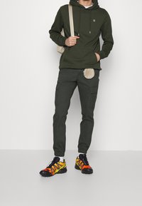 Hoodie verde scuro con logo, pantaloni cargo abbinati e scarpe multicolori con dettagli arancioni e gialli. Borsetta a cintura attaccata in vita.