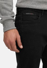 Hand ruht nahe der Hosentasche einer schwarzen Jeans, Person trägt graues Langarmshirt mit geripptem Bündchen.