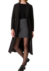 Manteau noir long avec revers crantés, deux boutons et fentes latérales, porté sur une jupe texturée grise et des chaussures noires, mettant en valeur une silhouette moderne.