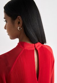 Crvena haljina s naboranim dizajnom i detaljem u obliku ključanice na leđima. Ima glatki ovratnik i teksturirani materijal koji poboljšava cjelokupni izgled.