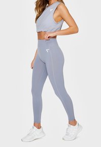 Jasnoszary crop top i legginsy z wysokim stanem z gładkiego materiału z dekoracyjnym szyciem. Noszone z białymi butami sportowymi.