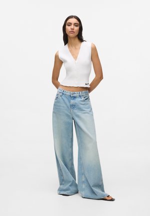 Woman wearing sleeveless white buttoned crop top and wide-leg light blue jeans with black sandals, standing against white background.