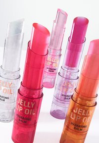 Fem Jelly Lip Oil-balsam i varierande höjder och färger: klar, rosa och orange, med transparenta cylindriska rör och metallisk text.