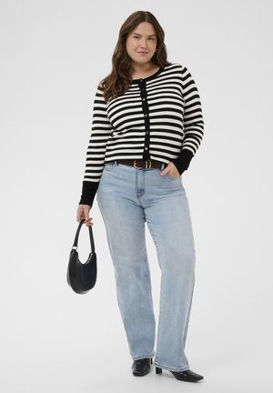 Woman wearing black and white striped buttoned cardigan, light blue jeans, black heeled shoes, holding a small black handbag with one hand.