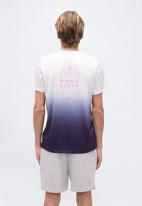 Άτομο με κοντά ξανθά μαλλιά φοράει αθλητική μπλούζα με διαβάθμιση από άσπρο σε σκούρο μπλε και ροζ κείμενο "MY SPORT IS HYROX", και ανοιχτό γκρι σορτς, στέκεται όρθιο.