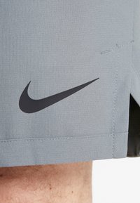 Nike-shorts i ljusgrått tyg har en svart swoosh-logotyp, platta sömmar och en texturerad yta som framhäver modern atletisk design.