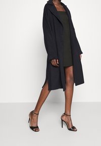 Long, dark navy coat made from a soft fabric, with a collar and wide sleeves, worn over a fitted olive green dress and black strappy heels.
