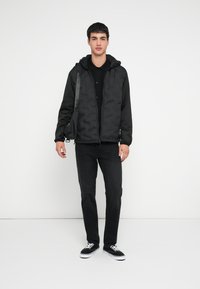 Sort boblejakke med glidelås og hette samt knappelukking, kombinert med svarte jeans og svarte joggesko. Klærne har en glatt tekstur.