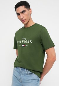 Zelené bavlněné tričko s kulatým výstřihem a krátkými rukávy. Na přední straně je bílé textové logo "Tommy HILFIGER 1985" nad malou grafiku vlajky.