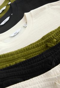Hauts à col rond en maille douce et pantalons taille élastique texturés en noir, blanc et vert olive, superposés et pliés avec des étiquettes de marque visibles.