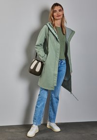 Veste imperméable verte clair avec capuche, intérieur gris, associée à un pull vert et un jean bleu. Accessoirisée avec un petit sac à main.