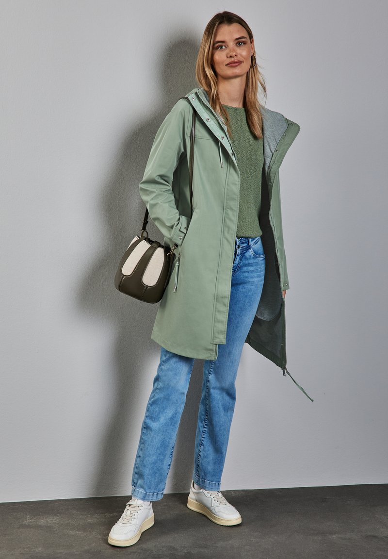 Veste imperméable verte clair avec capuche, intérieur gris, associée à un pull vert et un jean bleu. Accessoirisée avec un petit sac à main.