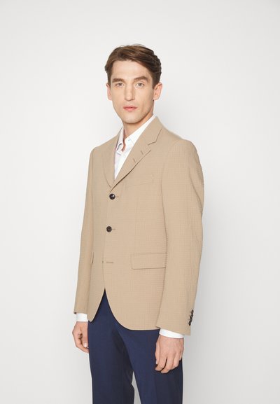 Blazer bege feito de tecido texturizado, com lapela em bico, dois botões frontais e um bolso no peito. Combinado com uma camisa branca e calças azul-marinho.