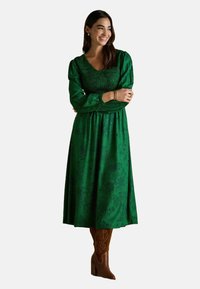 Žena sa spokojným úsmevom, prekrývajúca si ruky, oblečená v dlhých zelených kvetovaných šatách a hnedých vysokých podpätkoch, na jednoduchom pozadí.