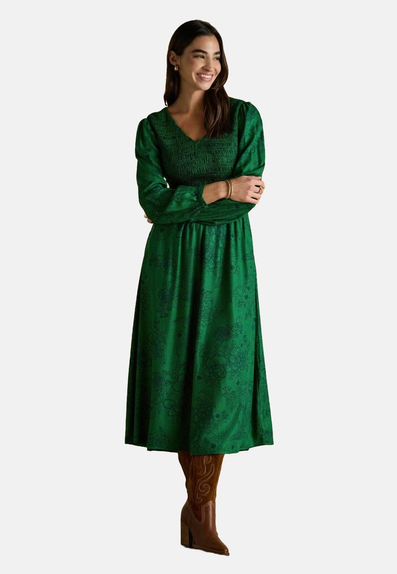 Žena sa spokojným úsmevom, prekrývajúca si ruky, oblečená v dlhých zelených kvetovaných šatách a hnedých vysokých podpätkoch, na jednoduchom pozadí.