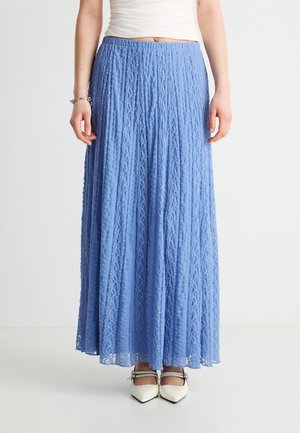 Γυναίκα που φοράει μακριά μπλε φούστα με υφή, λευκή αμάνικη μπλούζα, ασημένιο βραχιόλι και λευκά παπούτσια μυτερά με λουράκια αστραγάλου.