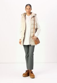 Gilet rembourré beige sur une chemise claire, associé à un pantalon vert olive et des chaussures plateforme marron à lacets, tenant un sac à bandoulière marron.