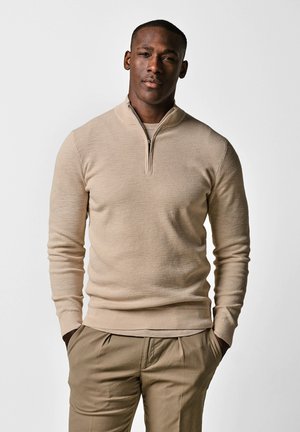Mann steht mit den Händen in den Taschen, trägt einen beigen Pullover mit Viertel-Reißverschluss und passende beige Hosen vor einem schlichten weißen Hintergrund.