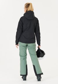 Schwarze wasserfeste Jacke mit Kapuze, kombiniert mit grünen Skihosen und schwarzen Skischuhen. Das Modell hält einen schwarzen Helm mit einer weißen Auskleidung.