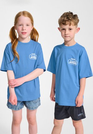 Två barn bär ljusblå kortärmade t-shirts med en logotyp. En bär denimshorts, den andra svarta shorts med ränder. Vit bakgrund.