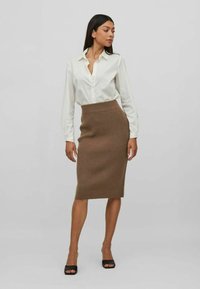 Camisa blanca de manga larga con botones, combinada con una falda marrón de canalé de longitud media, con una cintura ajustada y una textura suave. Zapatos de tacón abiertos de punta negra.