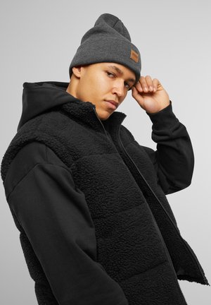 Gilet en polaire noir avec une fermeture éclair à l'avant, associé à un sweat à capuche noir. Bonnet tricot gris avec un patch en cuir marron.