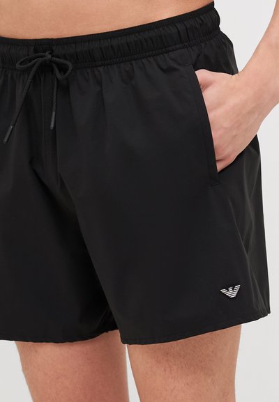 Bañador negro hecho de material ligero con cintura elástica, cordón, bolsillos laterales y un pequeño logo en el dobladillo inferior.
