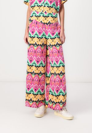 Pantalon à jambes larges avec un motif géométrique éclatant en rose, jaune, vert et noir, associé à des baskets blanches sur une personne debout.