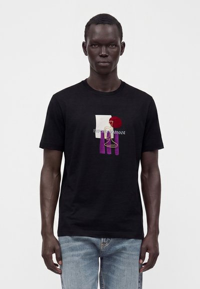 Άνδρας που φοράει μαύρο T-shirt Emporio Armani με πολύχρωμο αφηρημένο σχέδιο και ανοιχτό μπλε τζιν, στέκεται μπροστά σε λευκό φόντο.