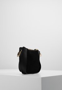 Malá černá semišová a kožená kabelka přes rameno se zlatým zipem a odnímatelnými klipy na popruh, vystavená na bílém podstavci na šedém pozadí.