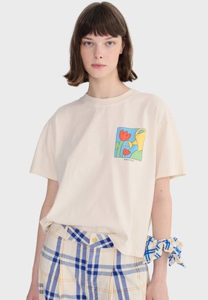 Jeune femme aux cheveux courts châtain, portant un t-shirt crème avec un motif coloré de fleur et d'arrosoir, associé à un pantalon à carreaux bleu et blanc.
