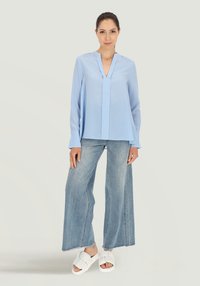 Jasnoniebieska bluzka z długim rękawem, w serek, z fałdą z przodu, połączona z dżinsami o szerokich nogawkach oraz białymi sandałami na płaskiej podeszwie. Gładka tekstura.