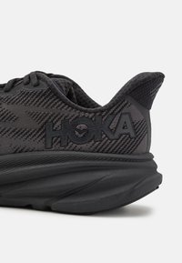 Scarpa sportiva nera con tomaia in rete, motivo testurizzato, tallone rinforzato e logo HOKA. Suola arrotondata con dettagli di ammortizzazione visibili.