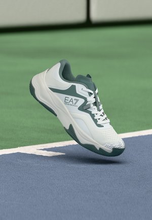 Chaussure de sport blanche et grise EA7 Emporio Armani flottant au-dessus d'une surface de court de tennis bleue et verte.