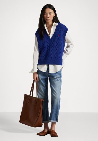Blue cable-knit sleeveless sweater over a white shirt, paired with light denim jeans and brown leather loafers, holding a brown tote bag.