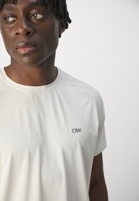 Camiseta deportiva de color gris claro con una tela texturizada, cuello redondo y mangas cortas. Presenta un logo negro en la parte izquierda del pecho.