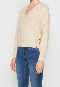 Beżowy sweter w stylu owijanym z fakturowanym wykończeniem, z długimi rękawami i bocznym wiązaniem. W połączeniu z niebieskimi dżinsami.