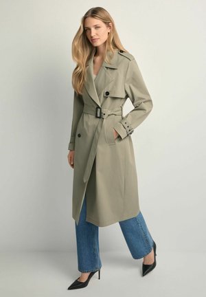 Kobieta o długich blond włosach, ubrana w beżowy płaszcz trencz z paskiem, niebieskie dżinsy i czarne szpilki, stoi na jednolitym jasnym tle.