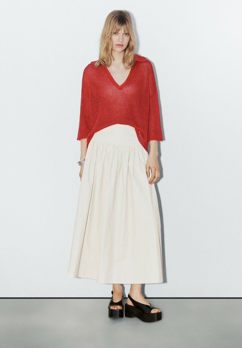 Rode gebreide top met V-hals en driekwartmouwen, gecombineerd met een witte geplooide maxi rok. Zwarte plateauschoenen maken de look compleet.