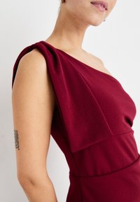 Burgunderett kjole med en skulder og draperte detaljer på skulderen, tettsittende silhuett og glatt tekstur. Designet for en skreddersydd passform.