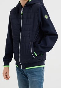 Navy modrá péřová bunda se zipem vpředu, zelené akcenty na kapuci a manžetách, s logem na rukávu a bočními kapsami.