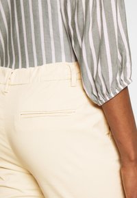 Närbild av en person i beige byxor med bakficka och en gråvit randig tröja med puffärmar.