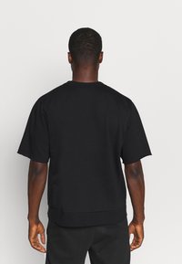 Hombre con cabello rizado corto, vistiendo una camiseta de manga corta negra lisa y pantalones negros, de pie frente a un fondo blanco liso.