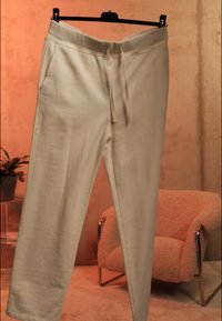 Φόρμες σε κρεμ χρώμα με κορδόνι στη μέση κρεμασμένες σε μαύρη κρεμάστρα μέσα σε εσωτερικό χώρο, με ροζ υφασμάτινη πολυθρόνα και φυτό στο φόντο.